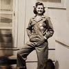 A woman in World War II-era work coveralls poses with one foot resting on a step.