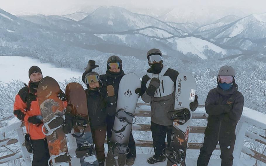 Five men holding snowboards pose for a picture in front of some mountains.