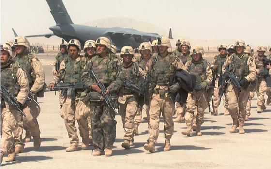 Soldiers in combat gear and carrying guns walk on a desert airstrip with an aircraft in the background.