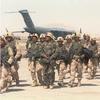 Soldiers in combat gear and carrying guns walk on a desert airstrip with an aircraft in the background.