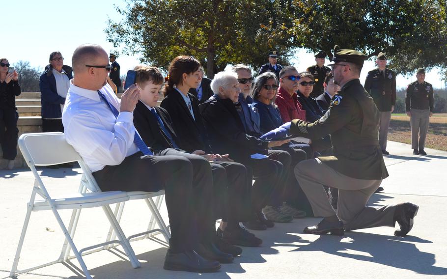 A soldier kneels in front of a group of people seated and hands an American flag to a woman.