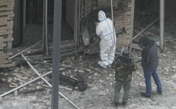 Investigators, one in a protective white suit, survey the site of a bombing, with bricks strewn along the ground.