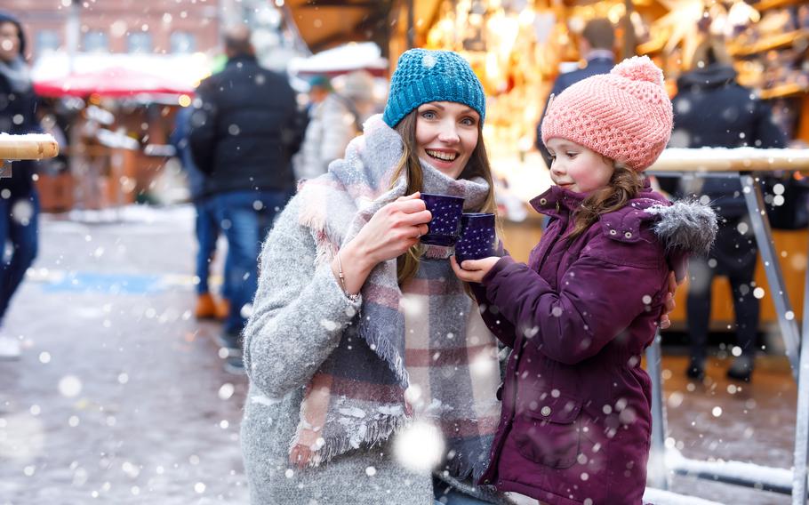 A woman and a young girl enjoy a drink in the show with the backdrop of a Christmas market.