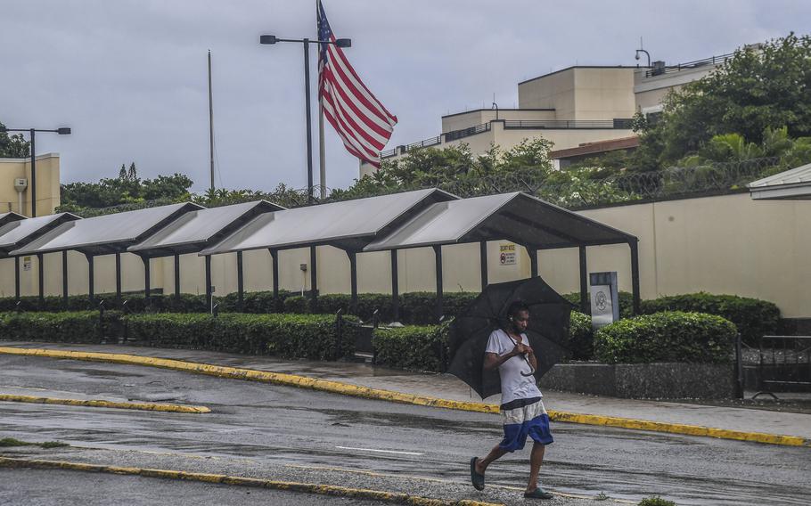 A man covers himself with an umbrella while he walks past a gated area with a raised U.S. flag.