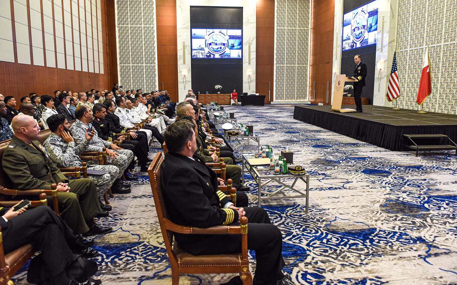 Rows of seated audience members watch a man speak on a slightly elevated stage.