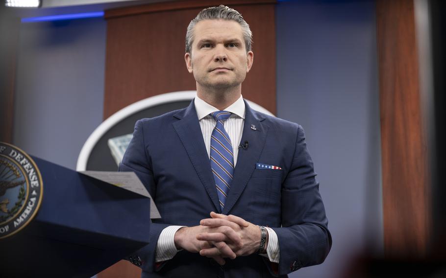 Pete Hegseth, wearing a dark blue suit, stands with his hands clasped in front of him in the Pentagon briefing room.