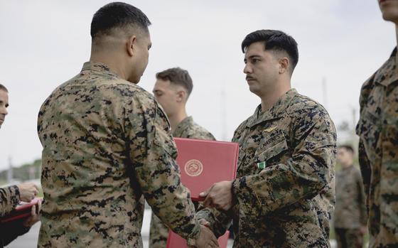 Two Marines shake hands as one hands the other a medal in a red portfolio case.