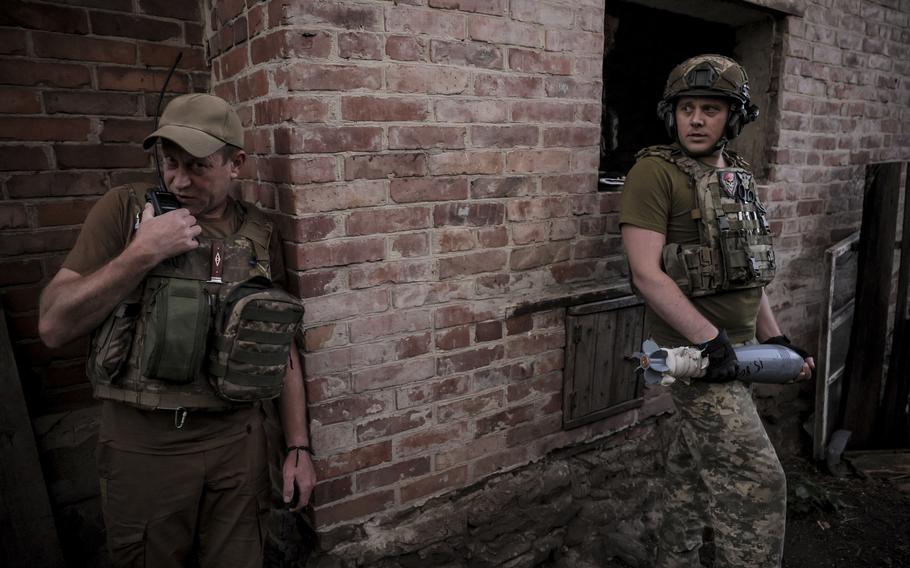 A Ukrainian soldier talks on a radio, and a second soldier holds a mortar round.