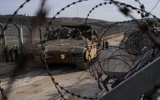 Soldiers, seen through coils of barbed wire stand next to an armored vehicle on a dirt road next to a fence.