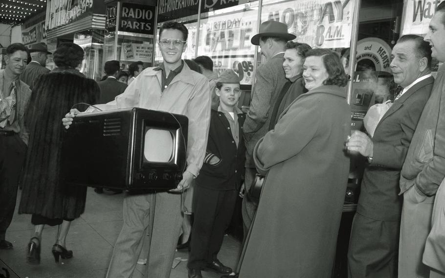 A black and white photo from 1954 of a man who just bought a new television set surrounded by other people waiting to get into a store.