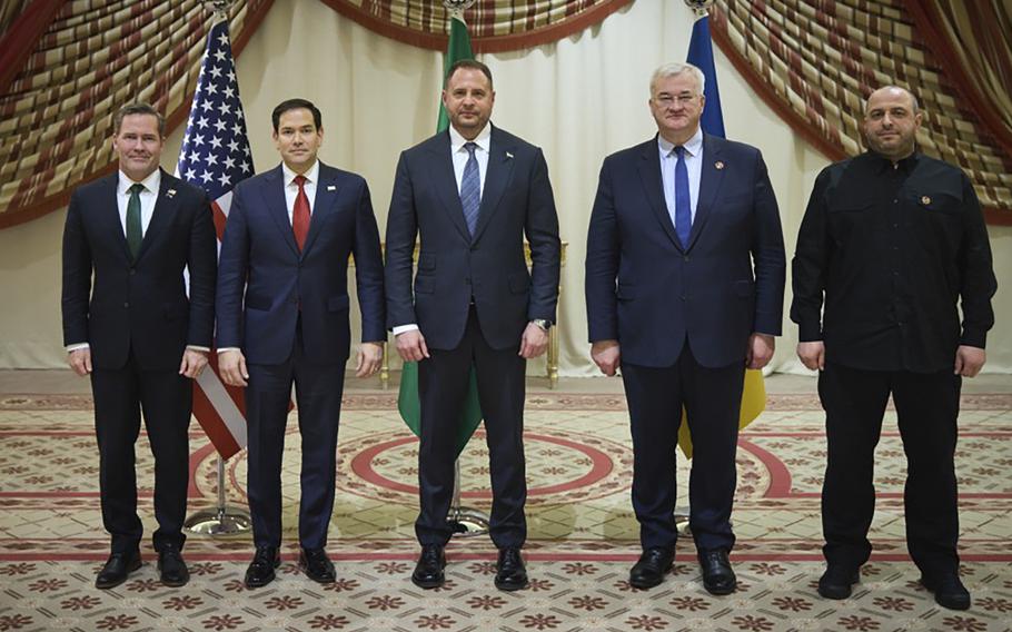 Five men in dark suits pose side-by-side in front of U.S., Saudi Arabian and Ukrainian flags.