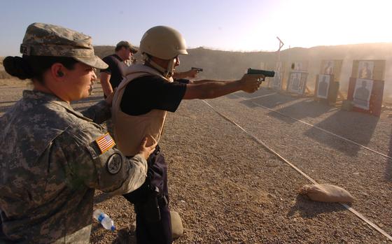 A man in a bulletproof vest and helmet fires a handgun on an outdoor shooting range while a military instructor stands behind him.