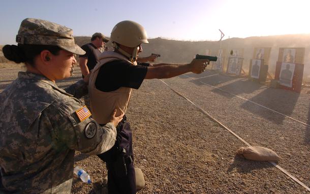 A man in a bulletproof vest and helmet fires a handgun on an outdoor shooting range while a military instructor stands behind him.