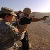 A man in a bulletproof vest and helmet fires a handgun on an outdoor shooting range while a military instructor stands behind him.