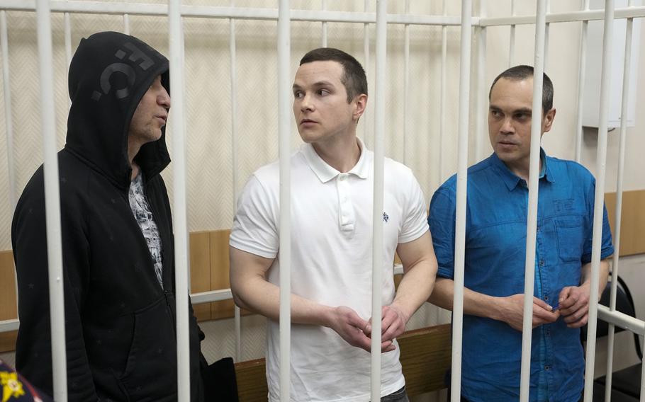 Three men in casual clothes are seen through the white bars of a cage.