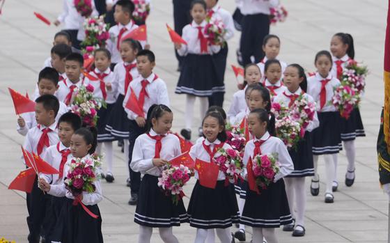 School children carrying Chinese flags and red and white bouquets walk together outside.