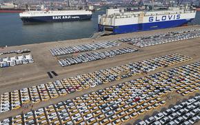 Cars are parked at a port with ships in the background.