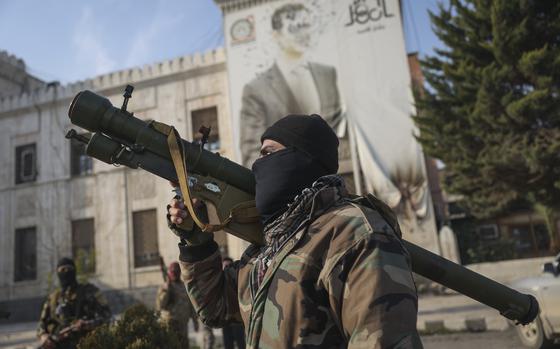 A Syrian opposition fighter holds a rocket launcher in front of the provincial government office, where an image of Syrian President Bashar Assad is riddled with bullets on the facade, in the aftermath of the opposition's takeover of Hama, Syria, Friday, Dec. 6, 2024. (AP Photo/Ghaith Alsayed)