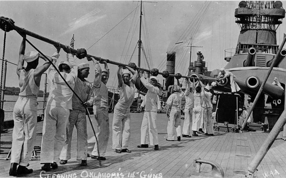 A black-and-white 1940s-era image of sailors in uniform standing in a line holding a large cleaning tool above theirs heads to push into the barrel of a gun turret.