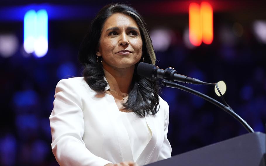 A woman with dark hair wearing a white suit stands at a podium and speaks into a microphone.