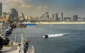 A boat travels towards a dock on the shore of a port with a city skyline in the background.