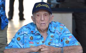 An elderly man is shown seated while wearing a blue shirt with U.S. flags and Navy ships and planes and a hat reading “Pearl Harbor Survivor.”