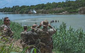 Troops watch from the river bank as people cross a river with a helicopter overhead.