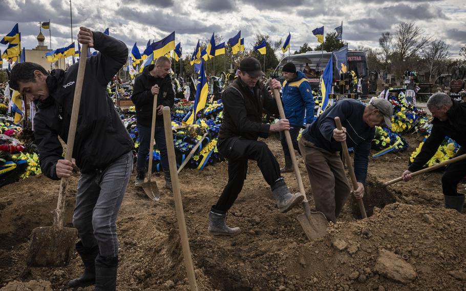 Ukrainian flags fly in the background as men with shovels dig graves.