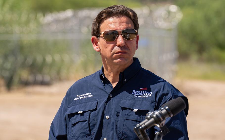 A man wearing a blue button down shirt and sunglasses stands outside in a desert climate area.