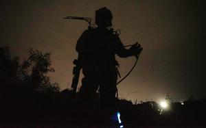 A military sevice member in tacitcal gear stands on a hillside while silhouetted against a backlit night sky.