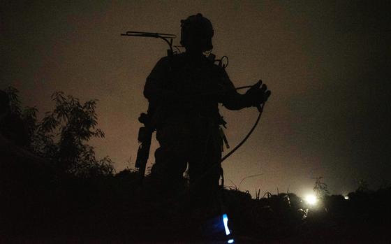 A military sevice member in tacitcal gear stands on a hillside while silhouetted against a backlit night sky.