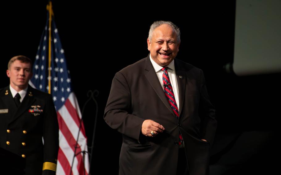 Del Toro smiles as he walks across a stage with a sailor and an American flag behind him.