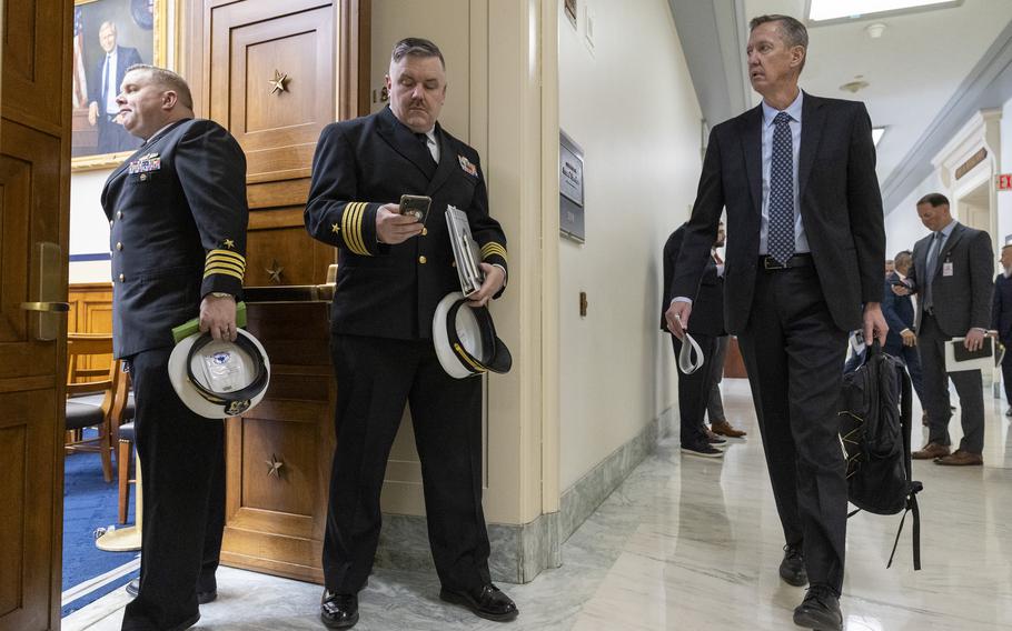 Two Navy officers in dress uniforms holding their hats stand in a doorway to a congressional hearing room as a man in a dark suit approaches from a hallway.