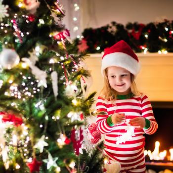 Happy little kid in red and green striped pajamas decorate Christmas tree in beautiful living room with traditional fire place. 