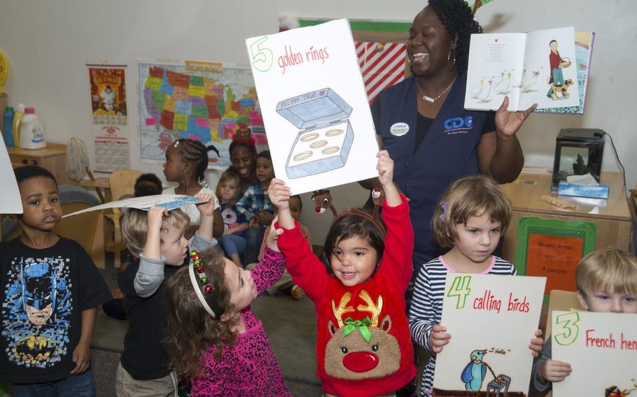 Children hold and their teacher hold signs depicting the 12 days of Christmas as they sing carols.