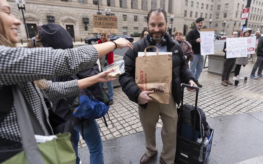 A man carries his belongings in a sack and rolling suitcase. Bystanders hold up signs thanking workers for their service.