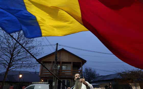 A Romanian flag covers the first third of the image as it is waved by a bearded man holding the flag pole in the bottom-center.