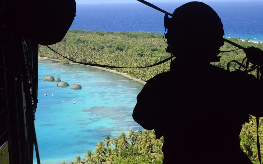 Boxes with parachutes attached float down toward an island in the distance as a service member watches from the back of the cargo bay.