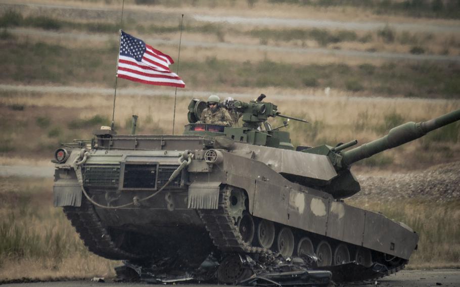 U.S. soldiers drive an M1A2 Abrams tank waving an American flag over a vehicle obstacle.