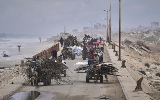 Displaced Palestinians in carts carry belongings and bundles of wood along a coastal road with a skyline of destroyed buildings in the background.