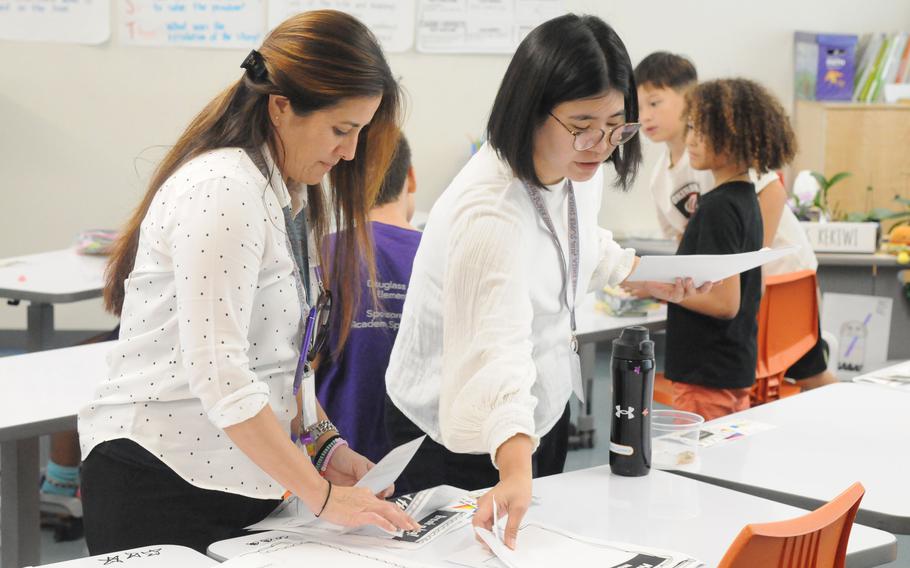 An American teacher and a Japanese teacher sort through papers on a tablet in a classroom.