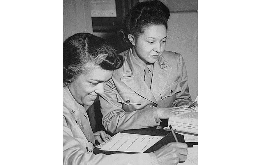 Two military women in WWII sit next to one another with papers and a pen, in a black and white photo.