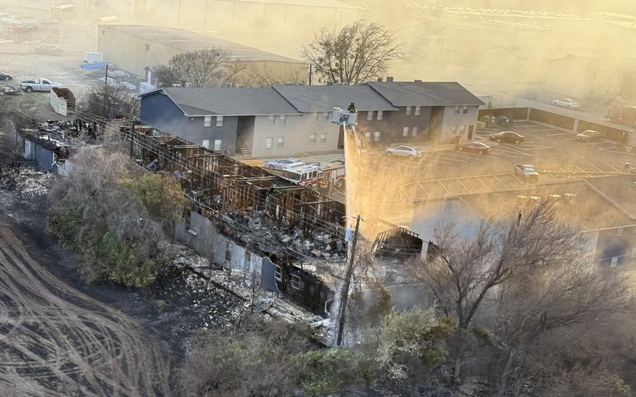 A firefighter in the bucket of a fire truck ladder, seen from above, uses a hose to spray water on the remains of a fire in an apartment complex.