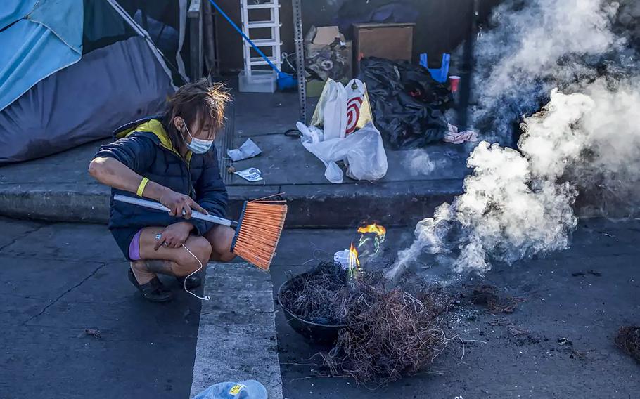 Toxic smoke billows up over a line of tents on the sidewalk as a person burns the plastic off copper wires in Skid Row, using a broom to fan the flames. 