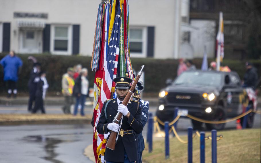 A man in a color guard uniform carrying a rifle leads a group carrying American flags.