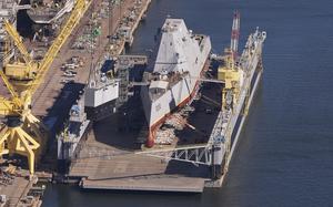 A stealth destroyer ship, seen from above, sits in dry dock at a shipyard.