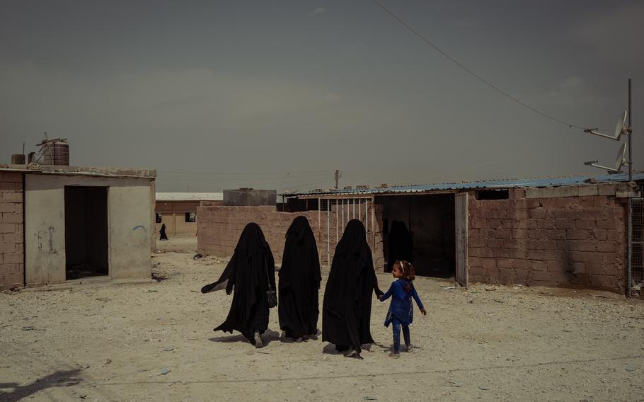 Three women on the left and a child on the right walk by one-story buildings.