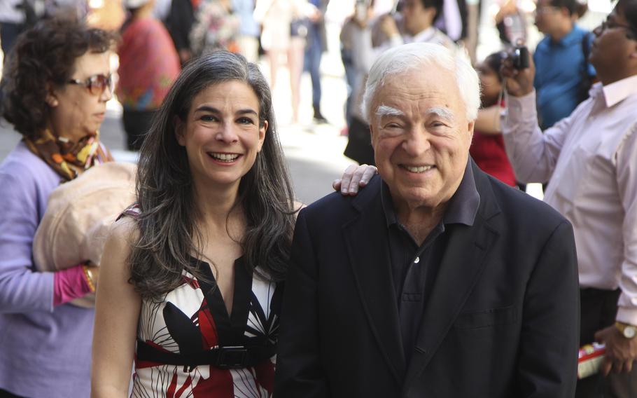 A woman and man stand smiling side by side with a crowd of people walking behind them.