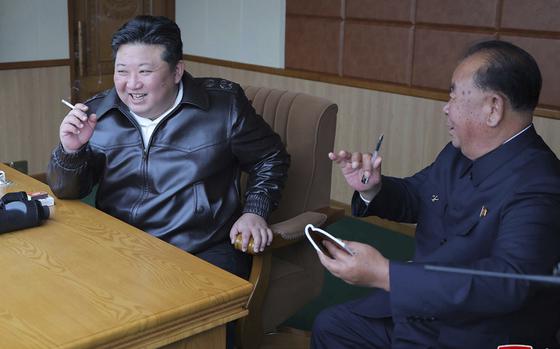 Kim Jong Un holds a cigarette while seated at a desk next to another seated official.