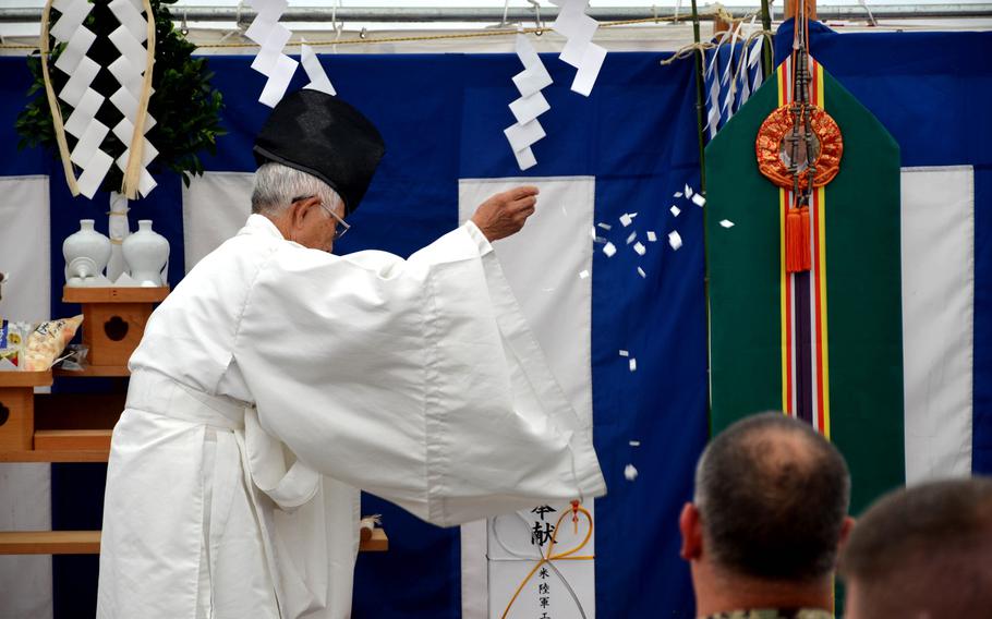 A Shinto priest in traditional clothing performs a ritual in front of a tapestry with religious symbols.